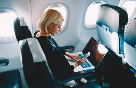 Blonde woman on in window seat on flight reading CTM News on laptop and looking at mobile